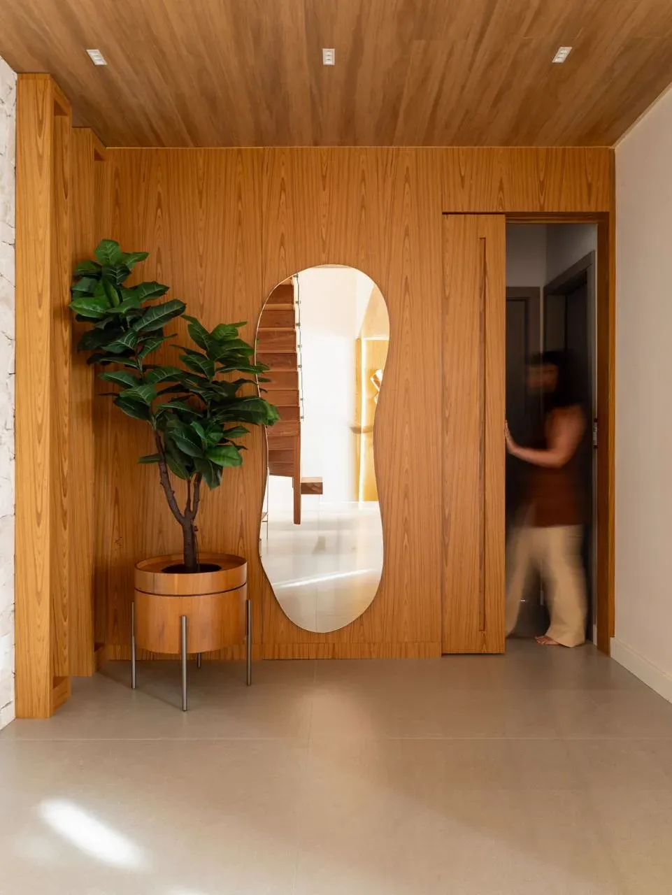 Hall de entrada revestido em marcenaria de madeira com espelho de formato orgânico e vaso alto com planta