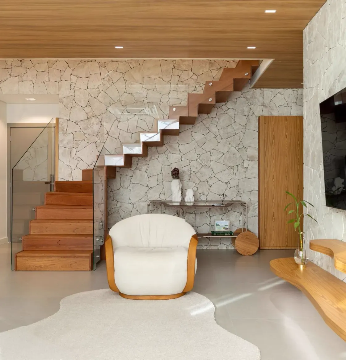 Hall residencial com escada de madeira maciça, parede revestida em pedra natural e poltrona bouclé off-white