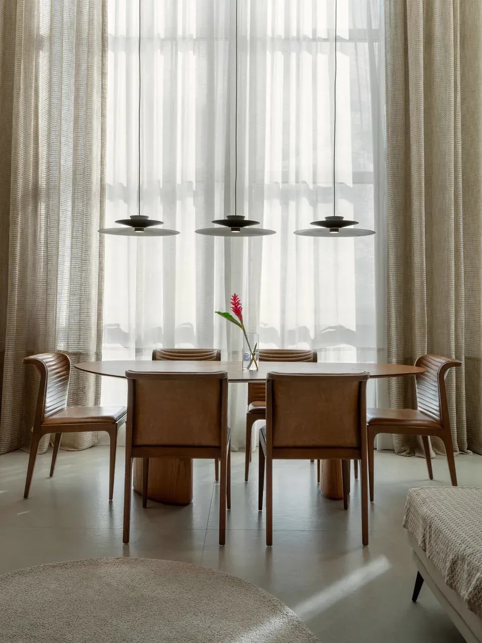 Sala de jantar com mesa oval de madeira, cadeiras de couro caramelo e três luminárias de disco diante de cortinas de linho